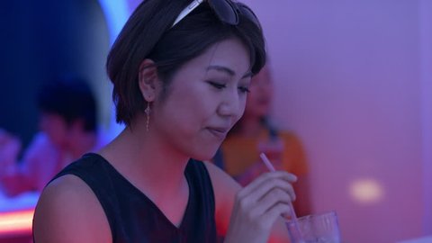 Japanese woman sitting with a cocktail and drinking it through a straw in a funky, cool bar with soft interior lighting. Close up shot on 4k RED camera.