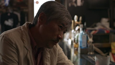 Tired, drunk Japanese man sitting at the bar counter with a cocktail in a dingy bar with soft day lighting. Medium shot on 4k RED camera.