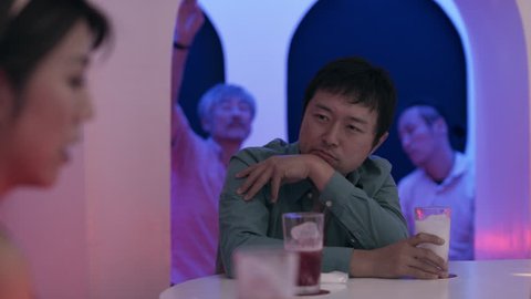 Pensive Japanese man drinking a cocktail and talking to a woman at the bar in a fun, cool bar with soft interior lighting. Medium shot on 4k RED camera on a gimbal.