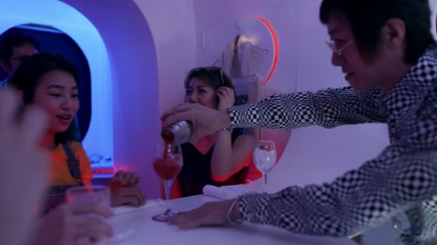 Fun Japanese bartender pouring a drink from a cocktail shaker into two glasses for two female customers in a funky, cool bar with soft interior lighting. Medium shot on 4k RED camera.