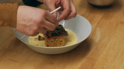 Professional chef finishing plating of sockeye salmon dish in a shallow bowl on a wooden counter in kitchen. Close up shot on 4k RED camera.