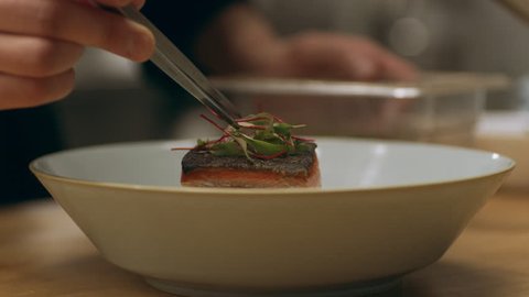 Focused professional chef carefully placing herbs on top of salmon dish in industrial kitchen with soft interior lighting. Close up shot on 4k RED Camera.