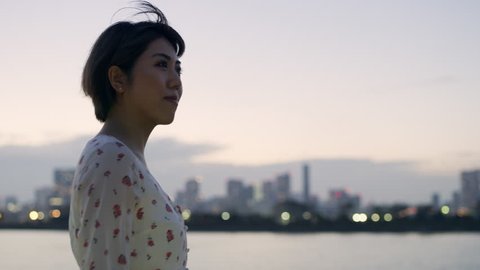 Smiling Japanese woman looking out at a skyline across the water with soft sunset lighting. Medium shot on 4k RED camera.