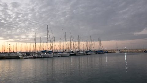 Peaceful view on small harbour with many parked sailing boats at early morning before sunrise. Seagulls flying above marina in front of blue sky with pink pastel clouds