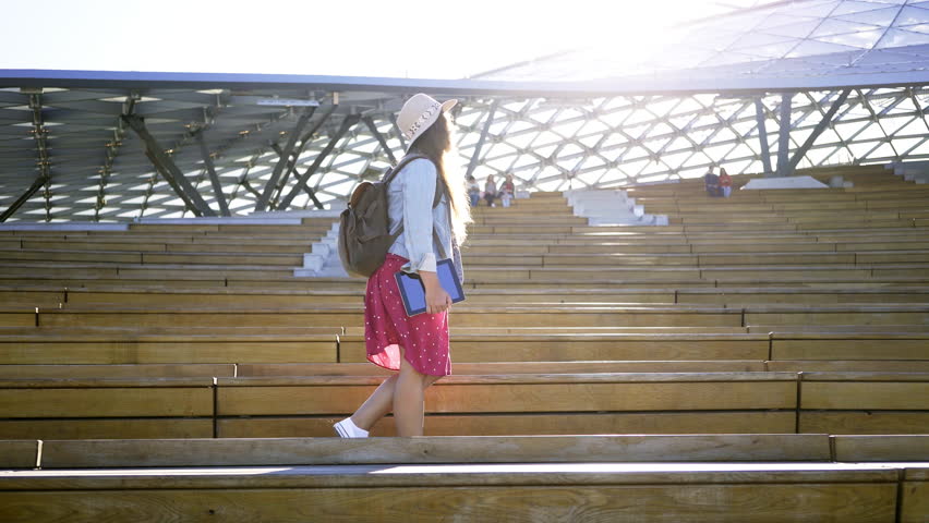 Sunny morning in modern park. Woman in denim and dress holding tablet and walking through wooden tribune