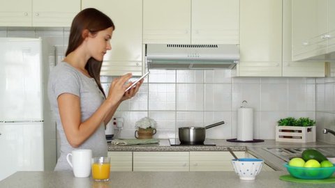 Young woman in pajamas enjoying early morning standing on kitchen drinking coffee or tea. Sleepy female using mobile phone reading news. Housewife browsing internet on cellphone