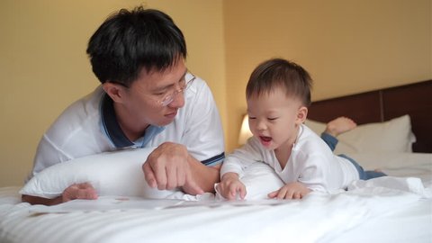 Smiling father and cute little Asian 2 years old toddler baby boy child reading bedtime story book, sitting on bed at home, Dad and son spending time together, Parents reading to child concept