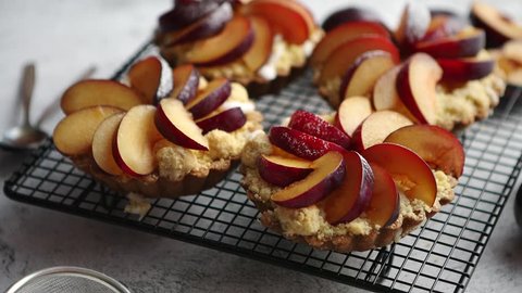 Close up of homemade crumble tarts with fresh plum slices placed on iron baking grill. Top lay on gray stone background with some whole plums, icing sugar, spoons on side