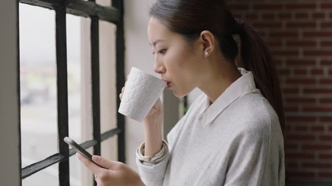 beautiful asian business woman drinking coffee at home using smartphone enjoying relaxed morning browsing messages looking out window planning ahead texting on mobile phone networking