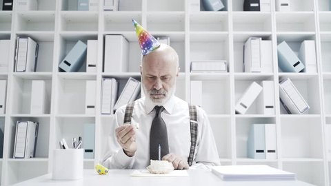 Sad businessman celebrating a lonely birthday in the office, he is blowing a candle on a small cake