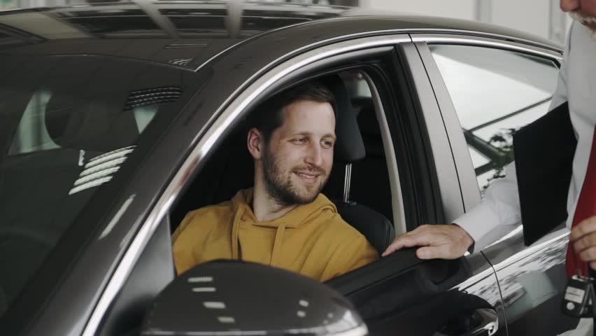 Young man, wearing casual closthing is getting the keys of his new car, from the sales man, Smiling at the camera.