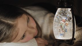 Cute girl lying on the floor watching snowflakes and lights in the Christmas globe lantern. Close-up video