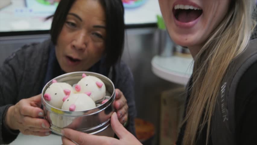 This slow motion video shows two diverse women tourists, an older asian woman and a young caucasian woman, enjoying cute pig shaped dim sum together at a restaurant in Hong Kong.