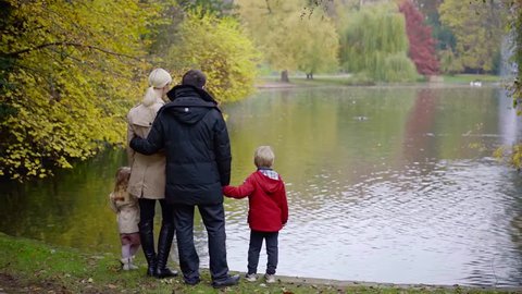 loving married pair is holding hands of little son and daughter standing on shore of small lake