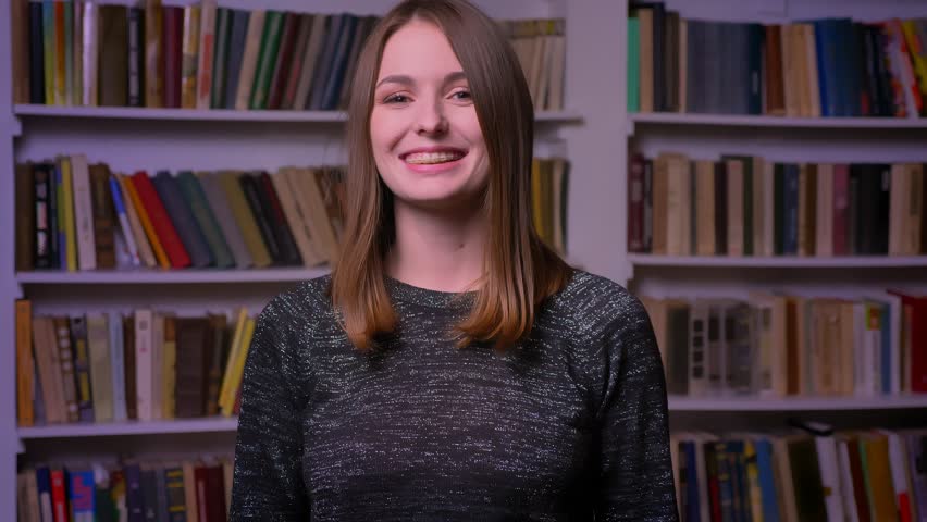 Redheaded caucasian girl in braces watches smilingly into camera, put her finger up to show like and respect on bookshelves background