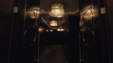 A steadicam shot through a hotel lobby with two elevators to a dark luxurious hallway. Beautiful chandeliers have warm muted light. Dark shiny doors are styled with geometrical metal details