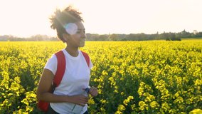 4K video clip of beautiful happy mixed race African American girl teenager female young woman hiking with red backpack and bottle of water in field of yellow flowers