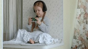 happy little girl with pigtails in headphones looks at the phone video, sits in a room on the windowsill, hid a blanket 4k