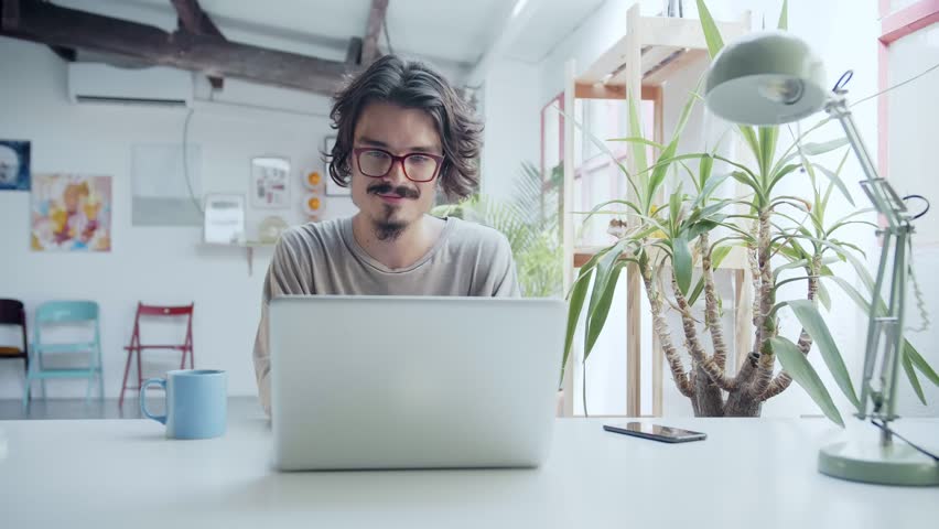 Young motivated hipster man with red glasses working reading on website or social network in computer or laptop next to a cup a tea or coffee in a bright modern loft or coworking space in the morning Royalty-Free Stock Footage #1020411811
