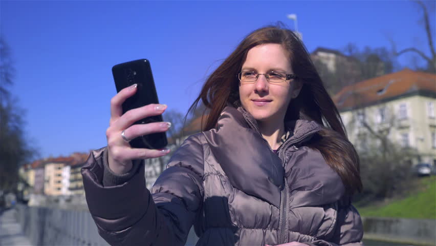 Taking a selfie with wind in hair HD. Woman in casual jacket holding smartphone mobile device in front and take a picture of herself in city center with castle in background.