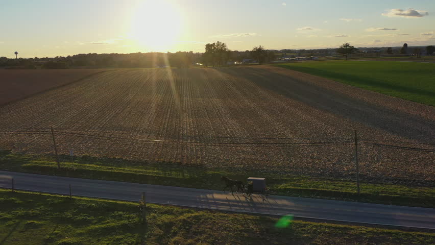 Aerial shot of a horse drawn carriage riding on an open stretch of road in Amish country during sunset