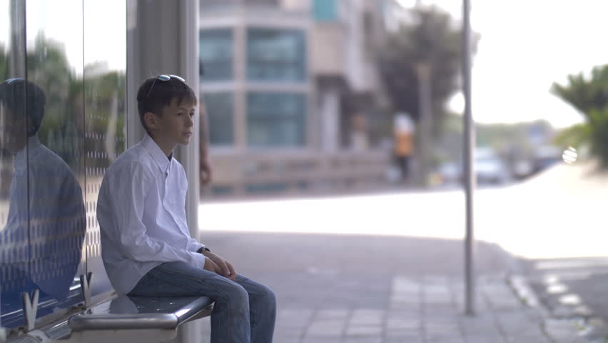 boy sits on the bus stop and waits for the bus, looks at watch Royalty-Free Stock Footage #1020455371