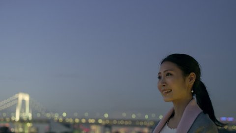 Happy beautiful Japanese woman looking out over the water to the skyline of buildings and a bridge with soft natural evening light. Medium shot on 4k RED camera.