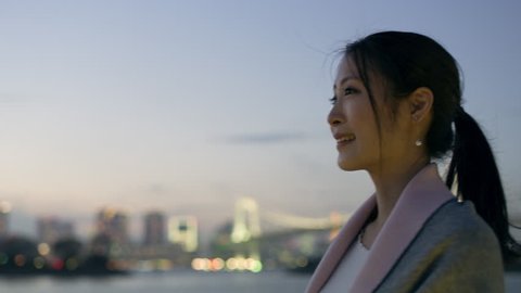 Smiling Japanese woman with ponytail looking out over the water to the skyline of buildings and a bridge with soft natural evening light. Medium shot on 4k RED camera.