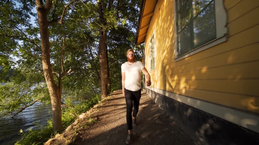 A tall Caucasian male with full beard walks by a canal in Stockholm Sweden. A sunny summer evening, green trees and blue sky. Steady glide cam footage. Nordic model with red beard.