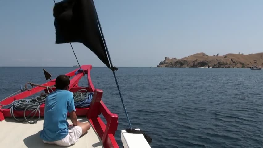 Man sits in moving boat with red bow of ship and black flag on background of sea surface and mountain islands on coastline under blue sky and white clouds in tropical nature of Bali Indonesia.