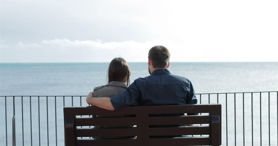 Back view of a couple in love hugging looking at ocean sitting on a bench on the beach