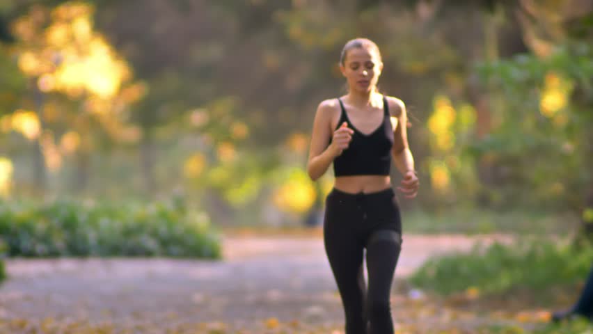 Young girl in jumpsuit with earphones running in autumnal park, stops to check the watch and continues jogging.