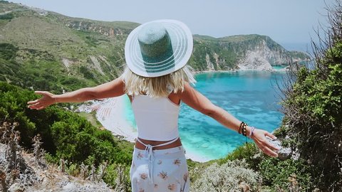 Woman with sun hat spreading arms in front of beautiful seascape on Kefalonia island. Blonde female tourist standing on hill admiring dreaming panorama of blue bay, arms wide open, Greece.