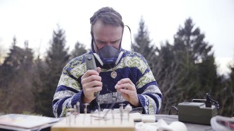Person with hand drill make a hole HD. Sliding over table with person wearing respirator mask and drilling a hole through a small pendant on table. Working outside in nature.