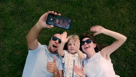 Top view of happy family outdoors spending time together. Father, mother and daughter are having fun and taking selfie on a smart phone while lying on a green floral grass.