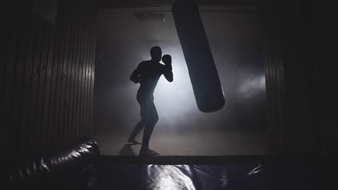Alone boxer hits punching bag in dark gym. Young man training indoors. Strong athlete in gym. Sport concept. Medium shot. Sportsman boxing in smoky studio