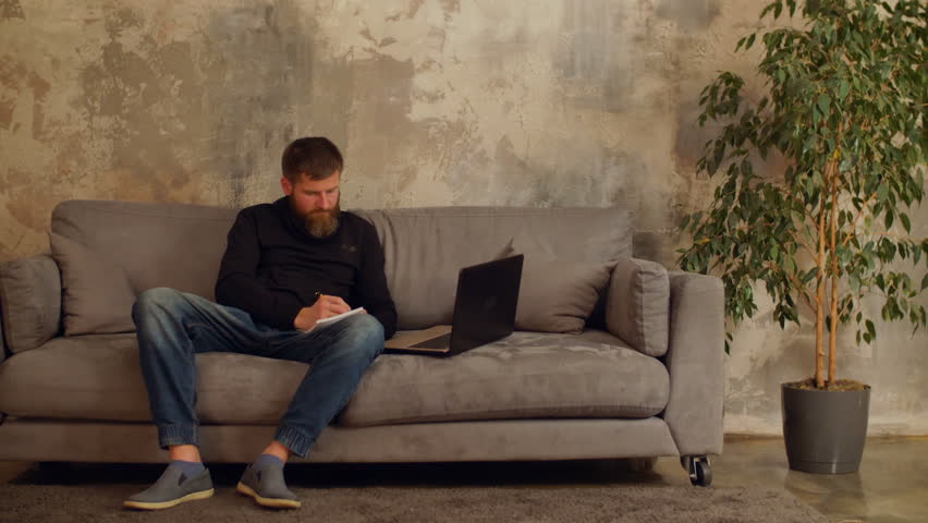 Busy bearded freelancer working on new business startup, networking online with laptop and writing notes while sitting on sofa at home. Handsome man with laptop pc creating new project in loft room.