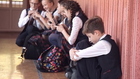 Selective focus of miserable schoolboy in uniform sitting alone on floor in school hallway with head on knees crying and feeling lonely, chatting and laughing classmates in background