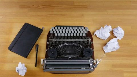 Typewriter, notebook and crumpled sheets of paper on a wooden table. Male hands type on a typewriter, and then throws out a sheet of paper.