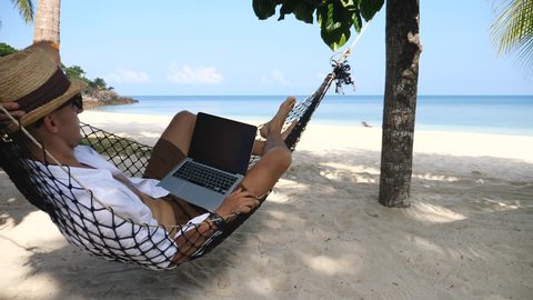 Man Working With Laptop Relaxing On Hammock On The Beach. Concept Of Digital Nomad, Remote Worker, Independent Location Entrepreneur.