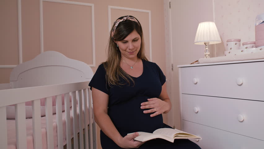 Pregnant Young Mom Sitting Reading A Book Caressing Belly, Baby Room Interior