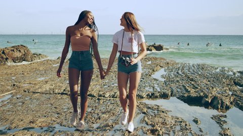Young lesbian couple hold hands and walk on coral rocks on a beach in sunny Gold Coast Australia. Wide shot on 4k RED camera.