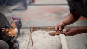 This close up video shows the hands of a man methodically putting burning incense on to an asian rooster shrine.