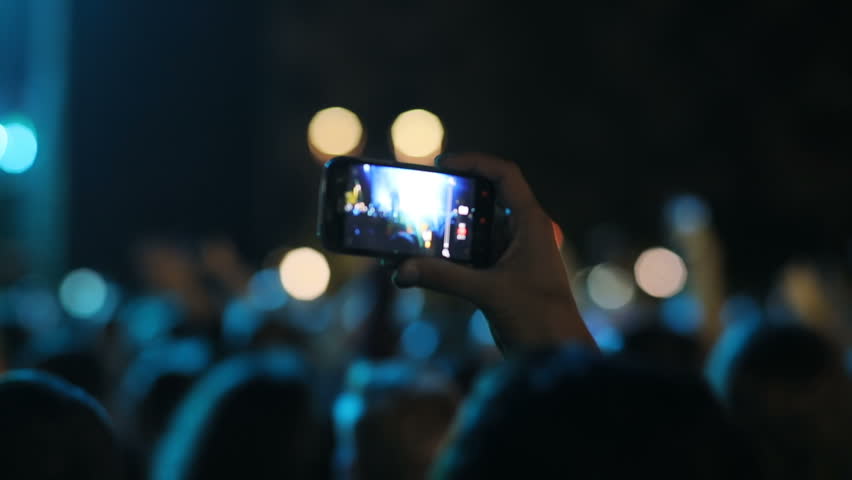 Back shot of person with mobile phone in hand. Fan making video at concert, using smartphone. Crowd. Event, outdoors.