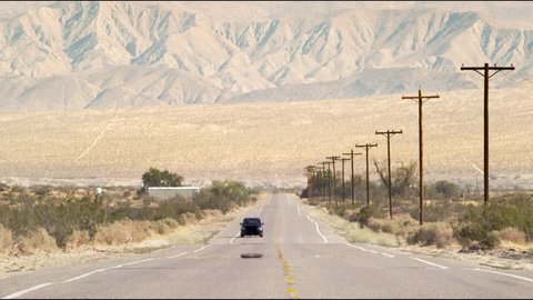 Long shot of a minivan driving down a hot desert road with mountains in the background.