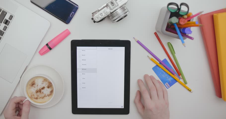 Top view of black tablet device lying vertically on the wooden office table. Male hands drink coffee and chat in tablet sitting at office table