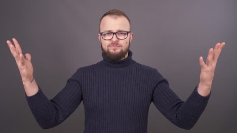 Portrait of young man with beard and glasses. Looking at camera with anger and waving his arms. Isolated on grey background.