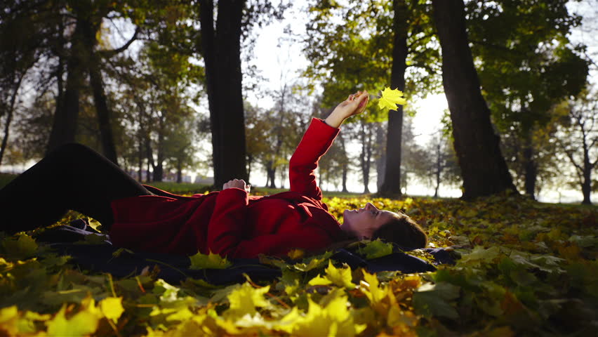 Woman play with leaf while lying on ground HD. Low angle view of attractive female person lying on autumn leaves and spinning one yellow leaf when sun shines through.