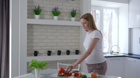 pregnant girl eats avocado and cuts vegetables with a knife in her hands standing at the kitchen table