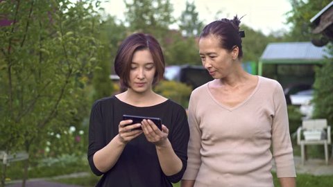 Asian middle aged woman and her young daughter walking in their garden and looking at photos on smartphone screen. Tracking real time medium shot